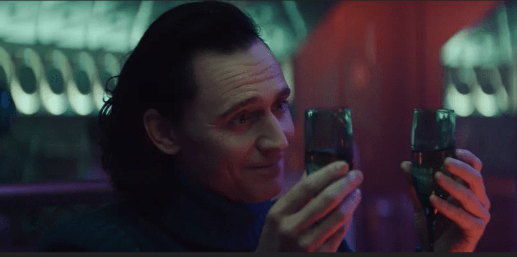 Loki holds up two glasses of champagne, smiling.