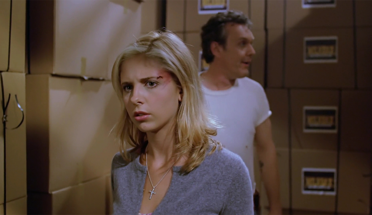 Buffy searches for Ethan with a serious expression. Giles stands out of focus behind her.